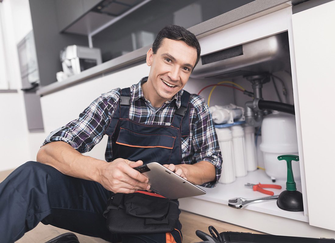 Business Insurance - Portrait of a Young Smiling Male Plumber Kneeling Next to a Kitchen Sink During a Job While Holding a Tablet in his Hands