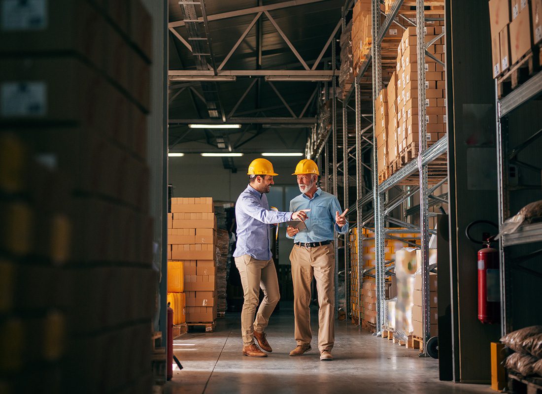 Safety and Risk Management - View of Two Employees Standing in a Warehouse Full of Products on the Shelves Wearing Yellow Safety Hats While Talking to Each Other