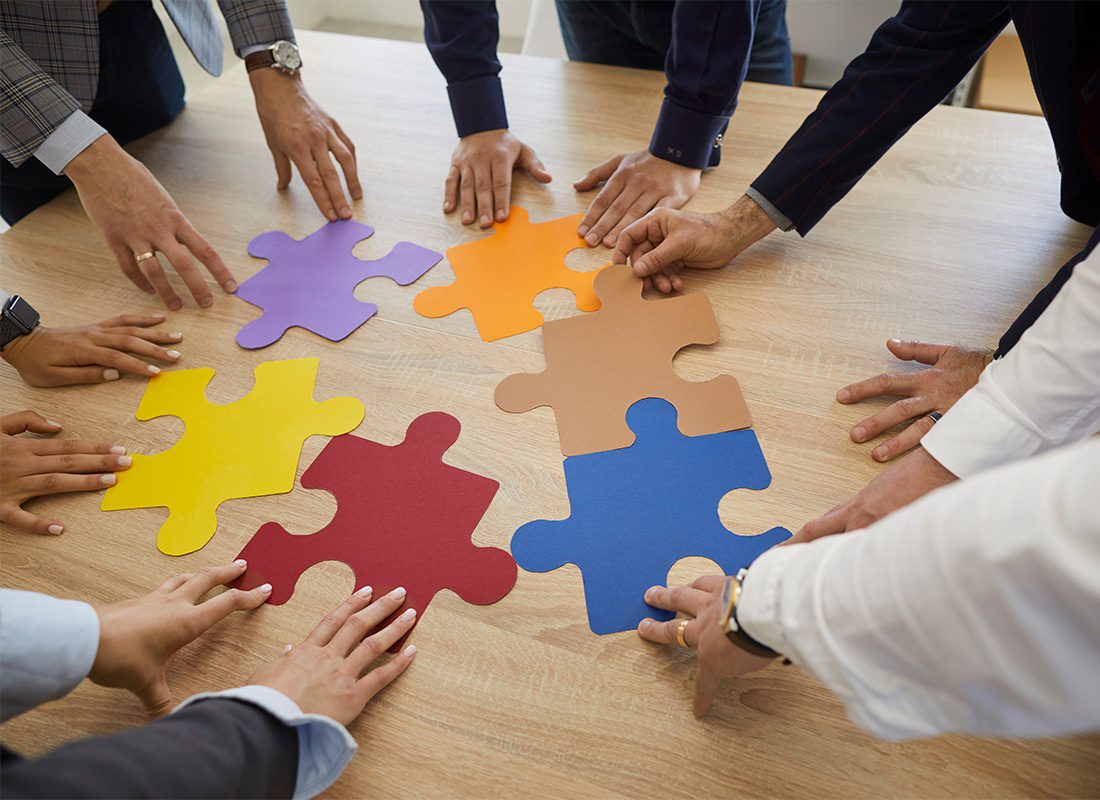 Insurance Solutions - Closeup View of the Hands of a Group of Business People Holding Out Colorful Puzzle Piece Cutouts on a Wooden Table During a Meeting