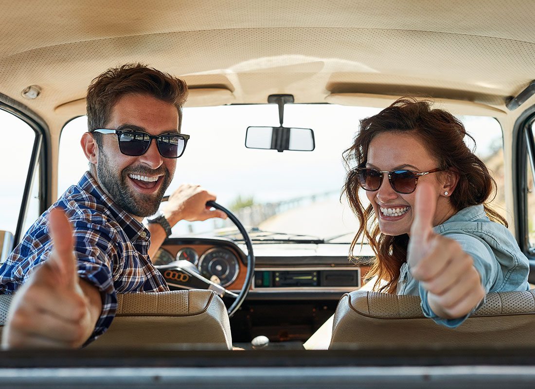 Review Us - Portrait of a Cheerful Young Couple Sitting in a Classic Car Looking Back and Giving Thumbs Up While on a Road Trip