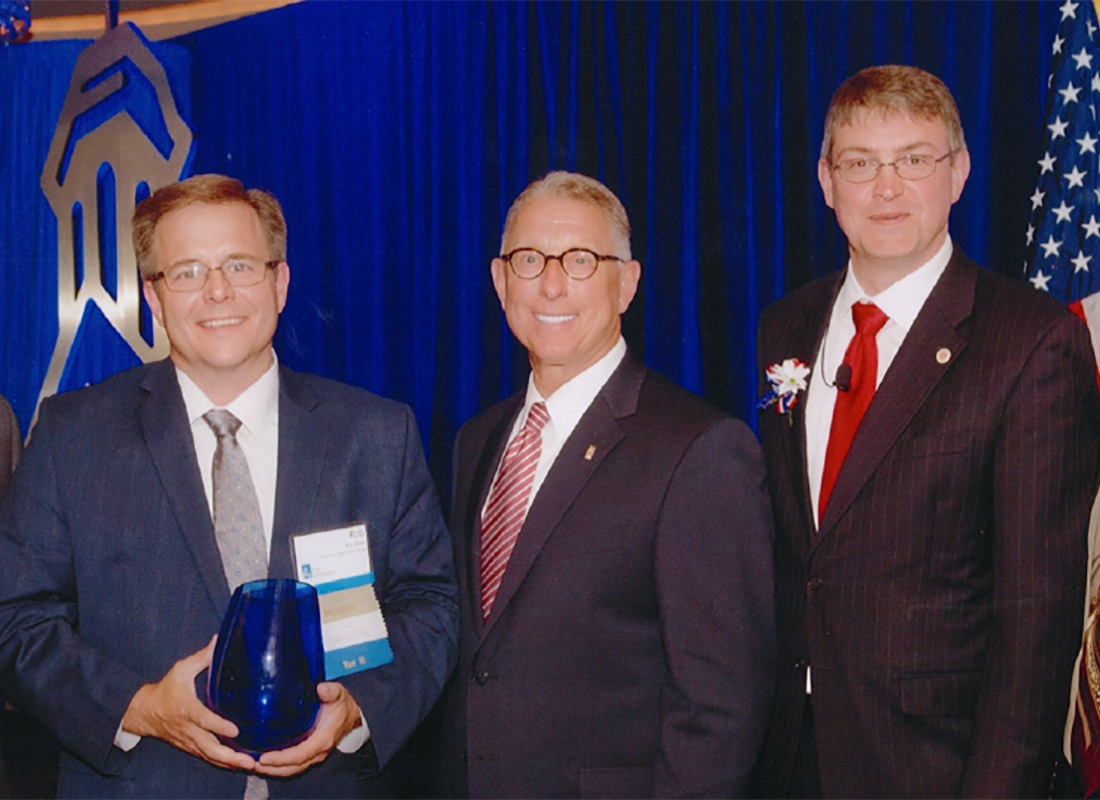 Our Awards - Covenant Cares President Robert Stone Holding an Award During an Award Ceremony While Standing Next to Two Business Men